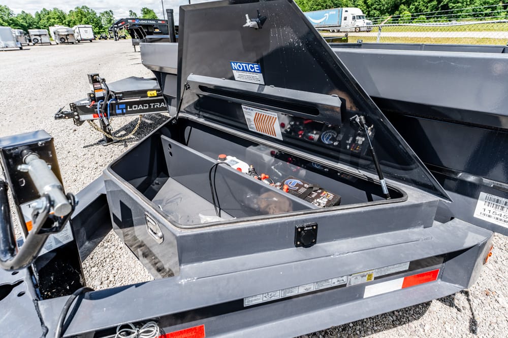 Open storage toolbox on a gray utility trailer, revealing a battery and control panel inside; other trailers parked on a gravel lot.
