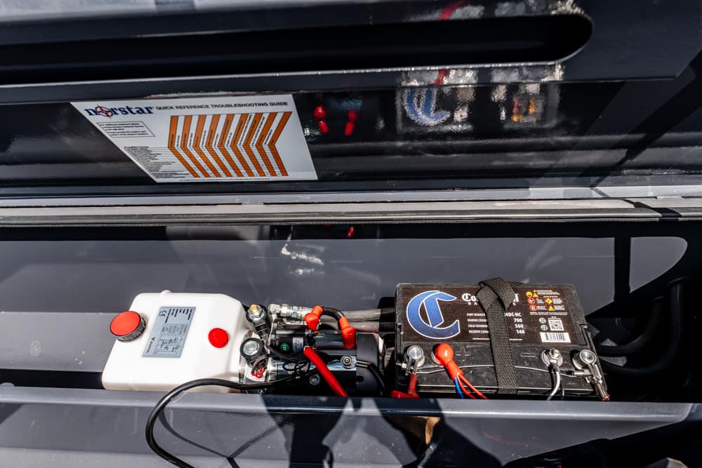 Open car hood showing a car battery with red/black jumper cables connected to a white portable jump starter/maintenance unit in the engine bay