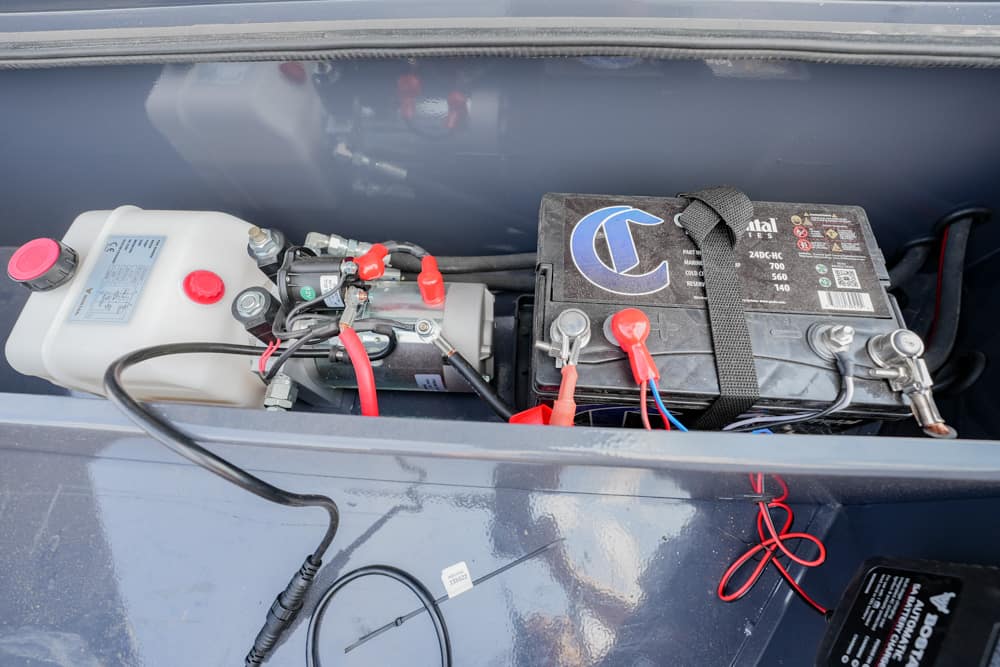 Battery and fluid reservoir in a boat storage compartment, with red/blue wires and a strapped battery connected to terminals.
