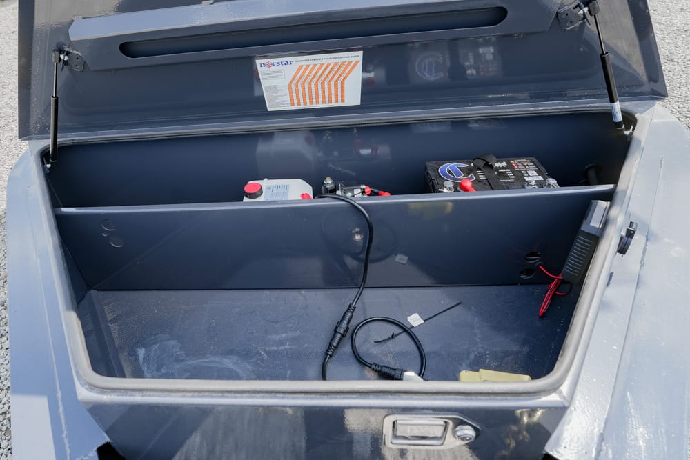 Open storage compartment with a car battery and red/black cables inside a blue metal bin under a lifted lid.