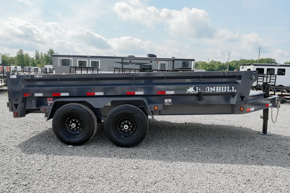 Dark gray dual-axle dump trailer in a gravel lot, labeled Iron Bull, with a front hitch and safety chains visible.