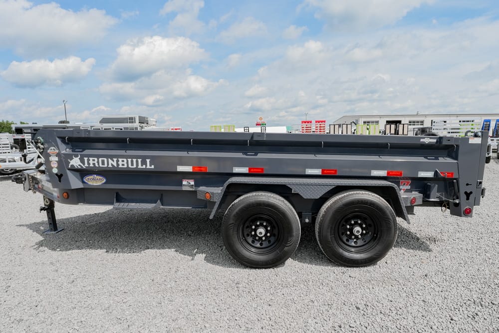 Side view of a dark gray Iron Bull dump trailer with dual axles parked on a gravel lot under a blue sky.