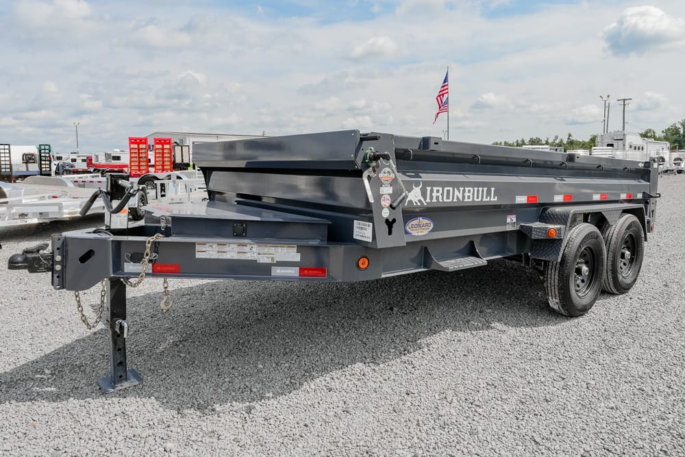 Dark gray Iron Bull dump trailer with dual axles on a gravel lot, American flag in the background, ready for use