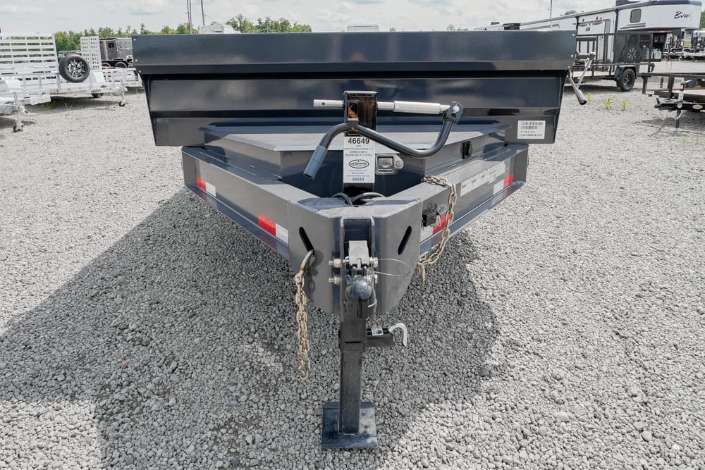 Rear view of a gray utility trailer on a gravel lot, showing the hitch, safety chains, and raised bed edge in front of other trailers.