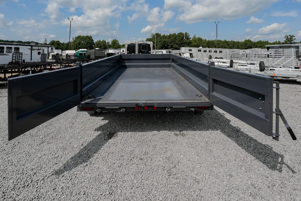 Rear view of a dark gray flatbed trailer with its side ramps opened wide, parked on a gravel lot among other trailers under a blue sky.