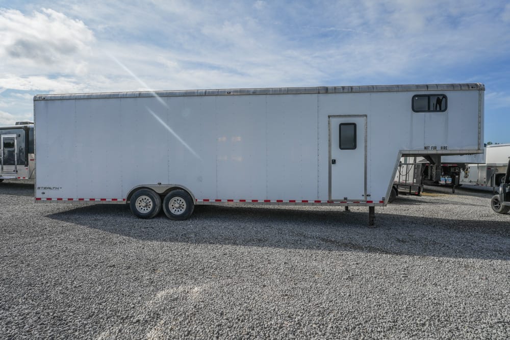 Long white cargo trailer with a side door and small window, two-axle wheels, parked on a gravel lot under a blue sky.