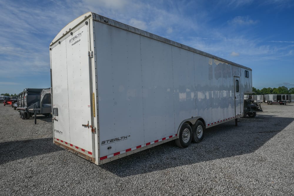 White enclosed cargo trailer with tandem axles parked on a gravel lot under a blue sky, with other trailers in the background.