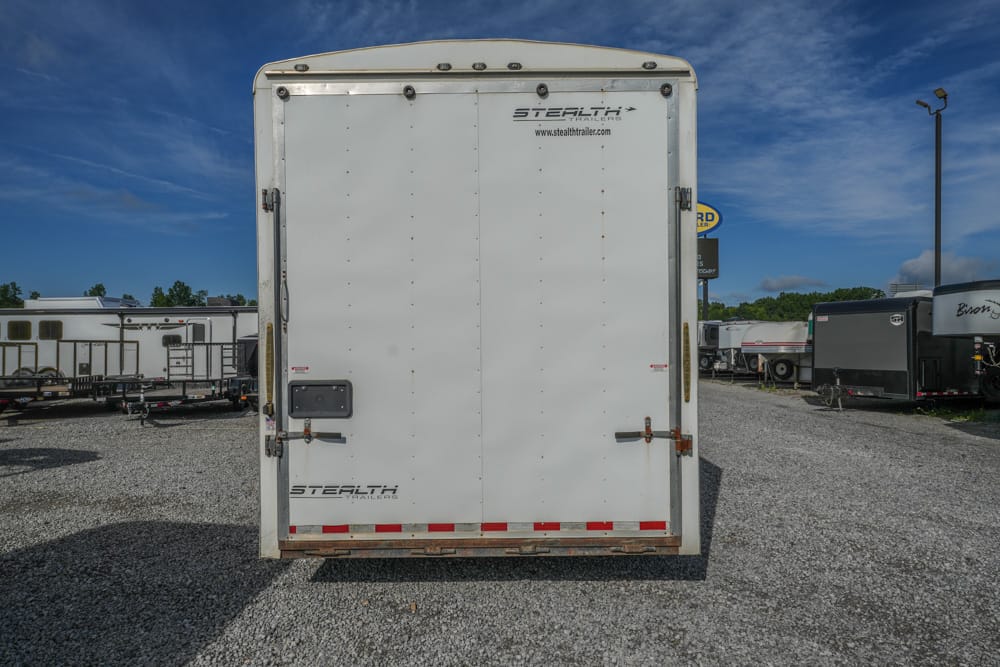 Rear view of a white cargo trailer with Stealth Trailers branding, parked in a gravel lot under a blue sky.