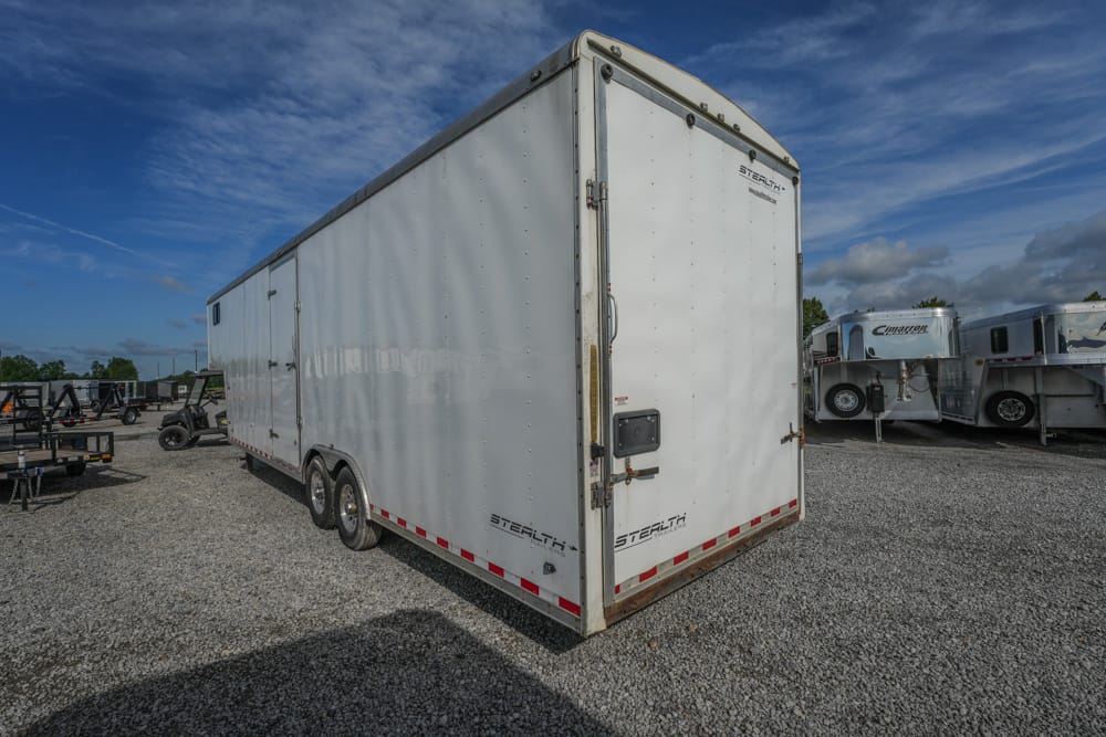 Large white enclosed cargo trailer with twin rear doors parked on a gravel lot, with other trailers in the background under a blue sky.