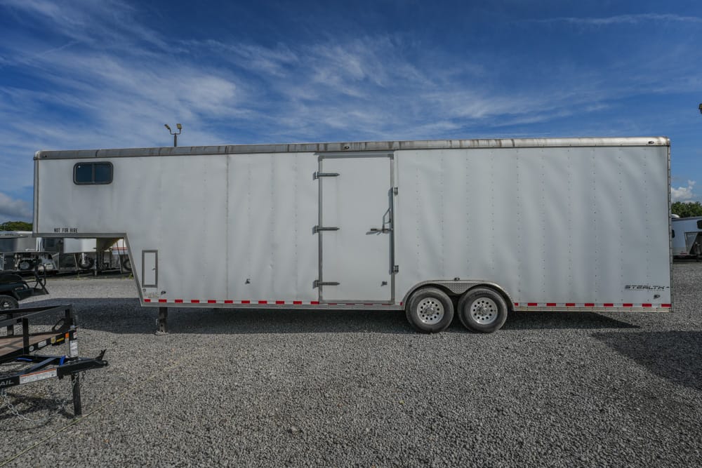 Large white enclosed cargo trailer with dual rear wheels parked on a gravel lot under a blue sky with wispy clouds.
