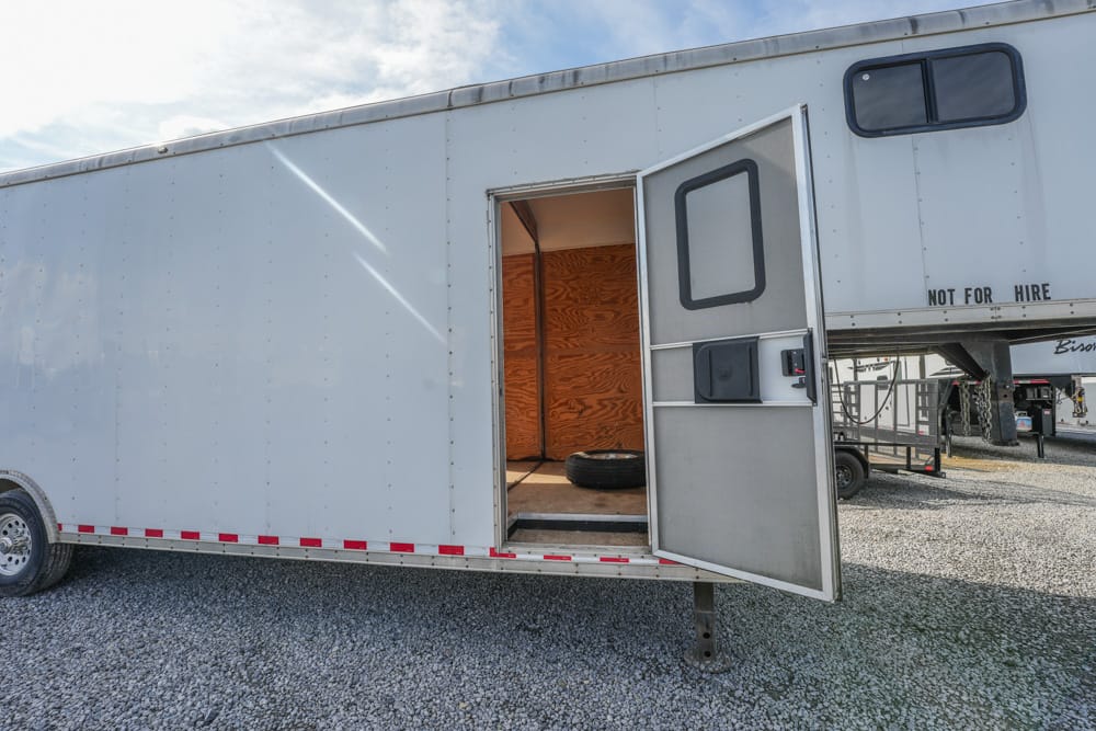 White cargo trailer with its side door open, exposing a wooden interior and a spare tire inside on a gravel lot