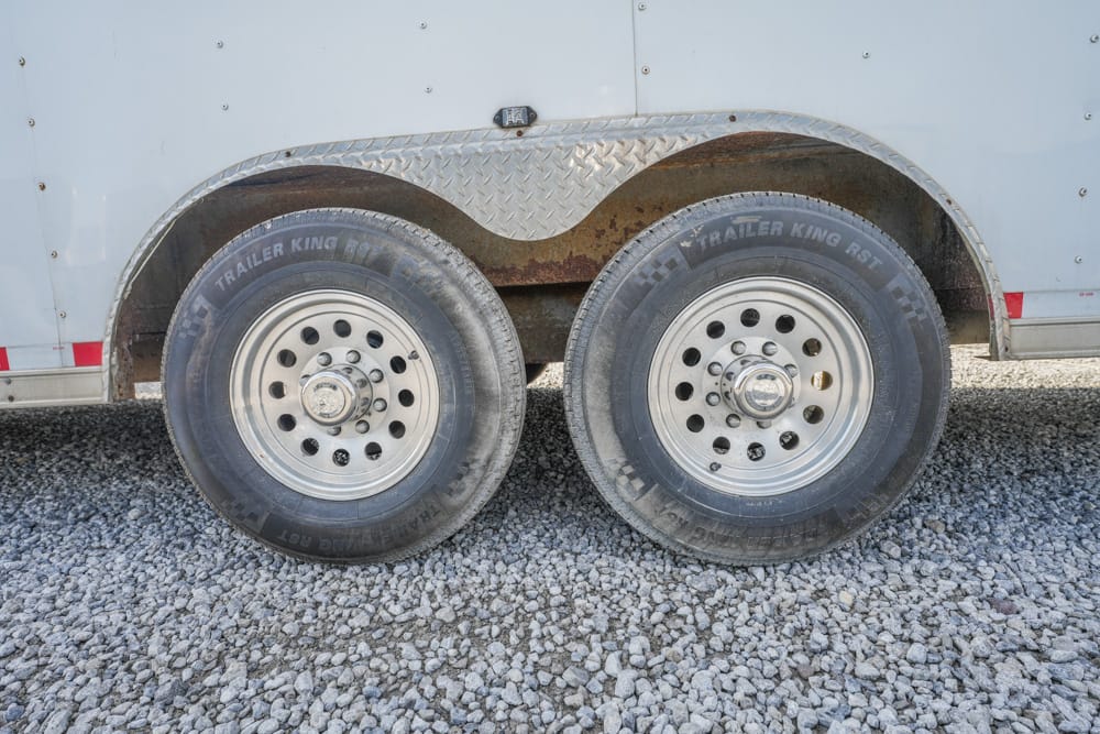 Dual trailer wheels with a diamond-plate fender and rust undercarriage, resting on a gravel surface.