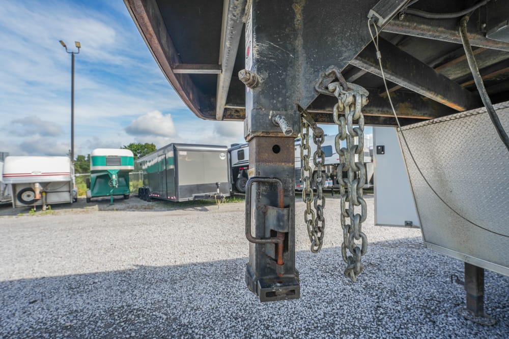 Close-up of a rusted trailer hitch with chains and a padlock under a flatbed trailer in a truck yard pool of trailers in the background.