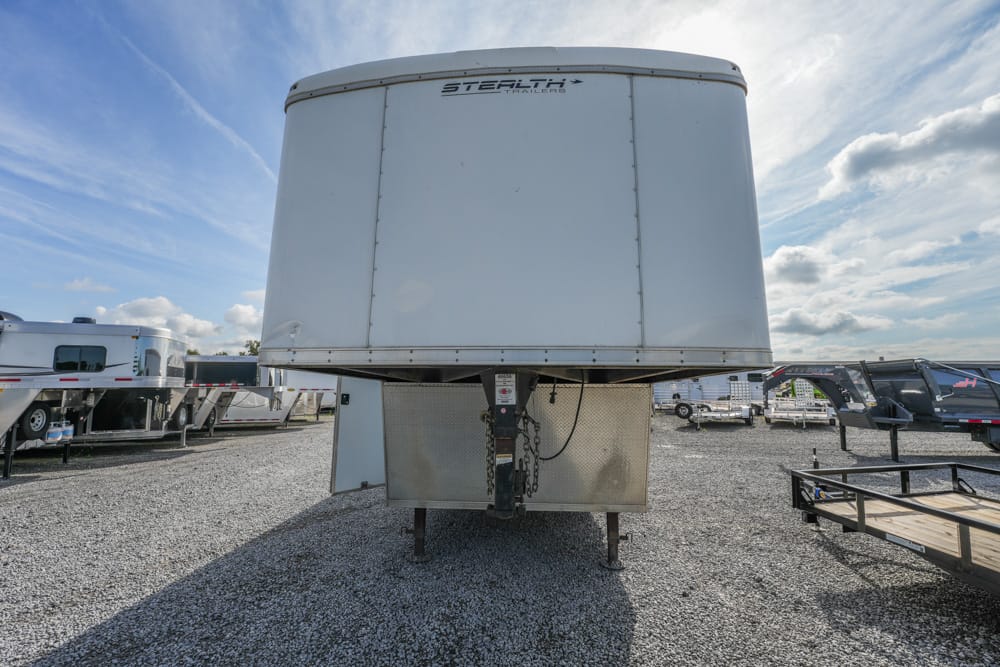 Front view of a tall white gooseneck trailer on a gravel lot, with other trailers and a blue sky in the background.
