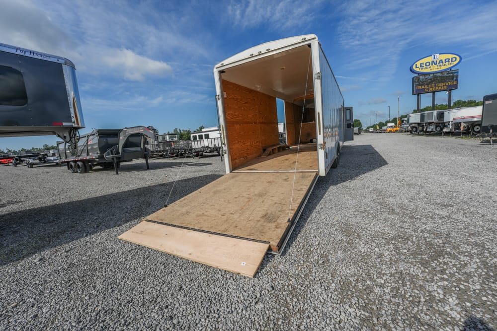 Open empty cargo trailer with ramp down in a gravel lot, other trailers nearby and a Leonard's Trailer Service sign in the distance.