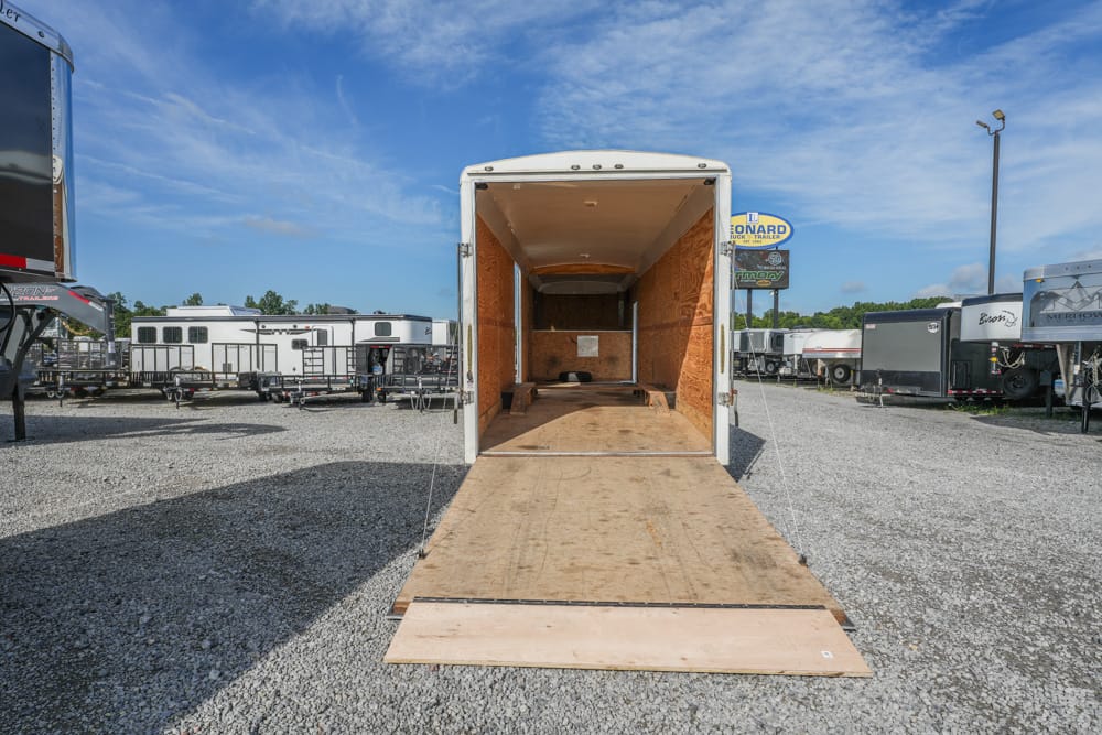 Open cargo trailer with plywood interior and ramp lowered for loading, parked in a gravel lot among other trailers on a sunny day