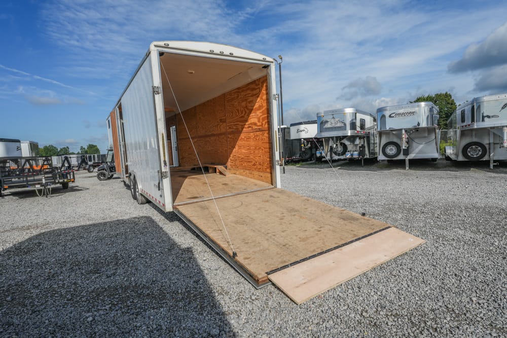 Open back of a large white cargo trailer with its wooden ramp lowered, parked in a gravel lot with other horse trailers nearby.