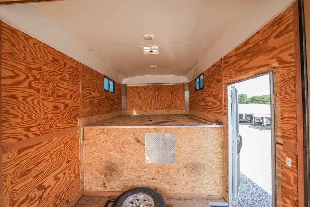 Interior of an empty wooden utility trailer with plywood walls and small side windows, a raised rear platform, and a spare tire on the floor near the open door.”,