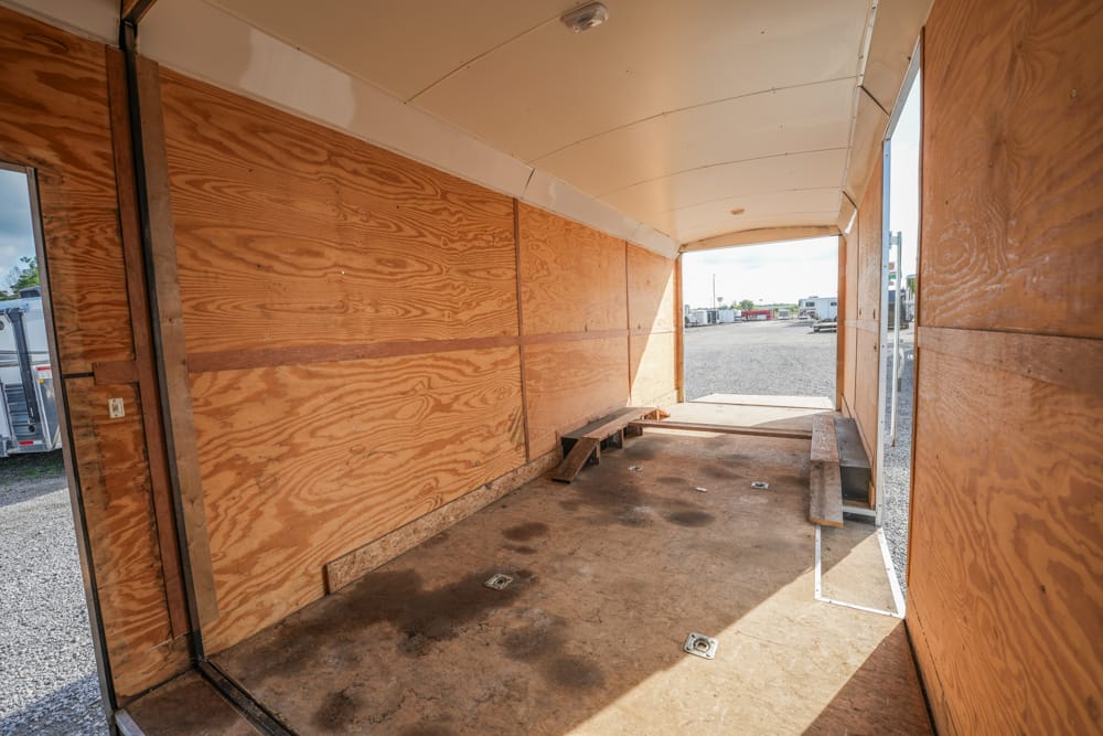 Interior of an empty wooden trailer with benches along the sides and an open rear doorway to a gravel lot outside.