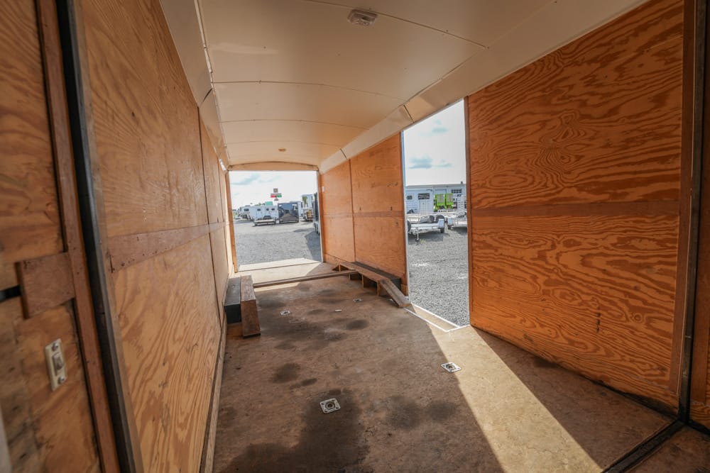 Interior of an empty wooden trailer with open rear doors, revealing a gravel lot and parked trucks beyond.
