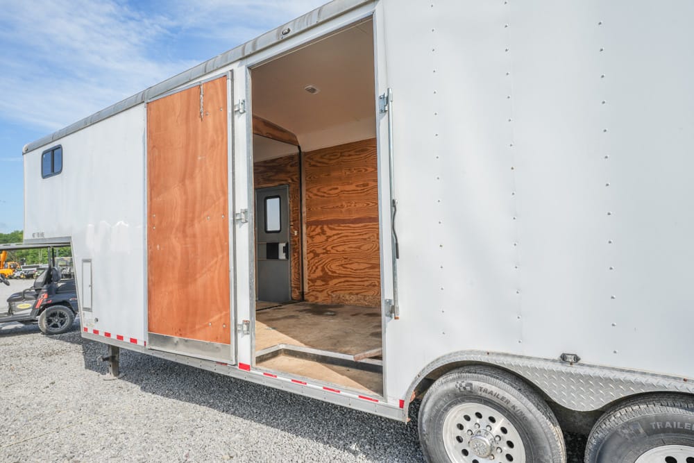 White enclosed trailer with rear doors open, revealing an empty cargo space and plywood interior on a gravel lot.