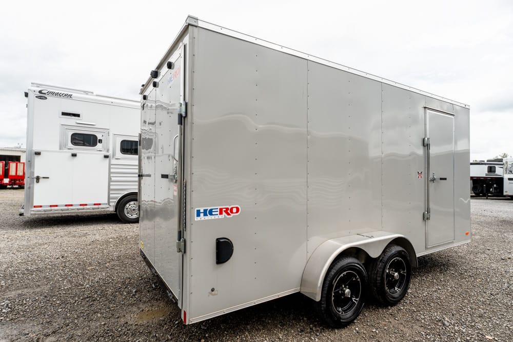 Gray enclosed cargo trailer with two axles and a side door, parked on a gravel lot; HERO logo on the side panel.