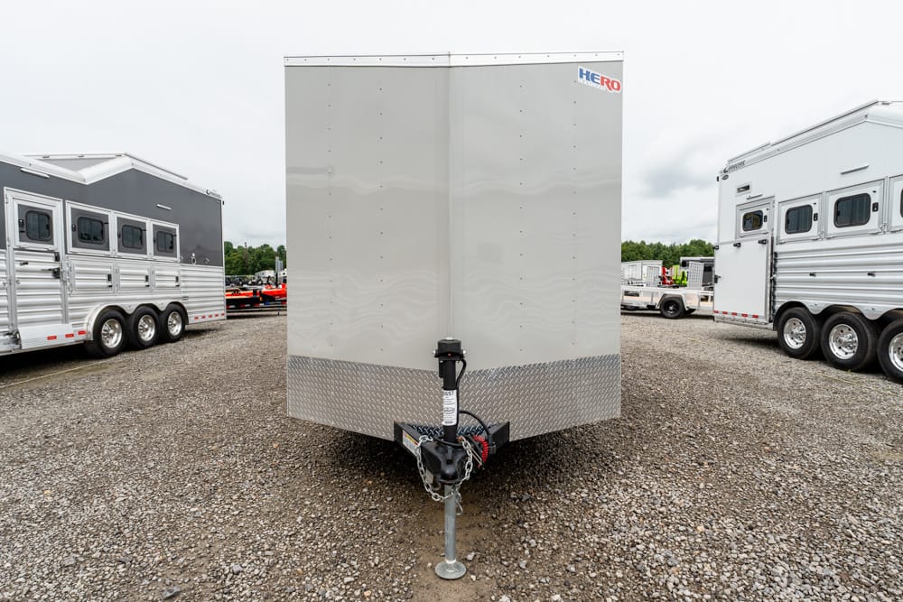Front view of a silver enclosed cargo trailer on a gravel lot, with its tongue jack and safety chains visible, diamond-plate trim at the bottom, and other trailers in the background.