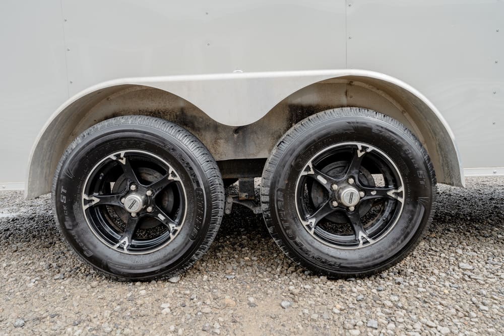 Tandem trailer with two black alloy wheels and a curved metal fender above them on a gravel surface.