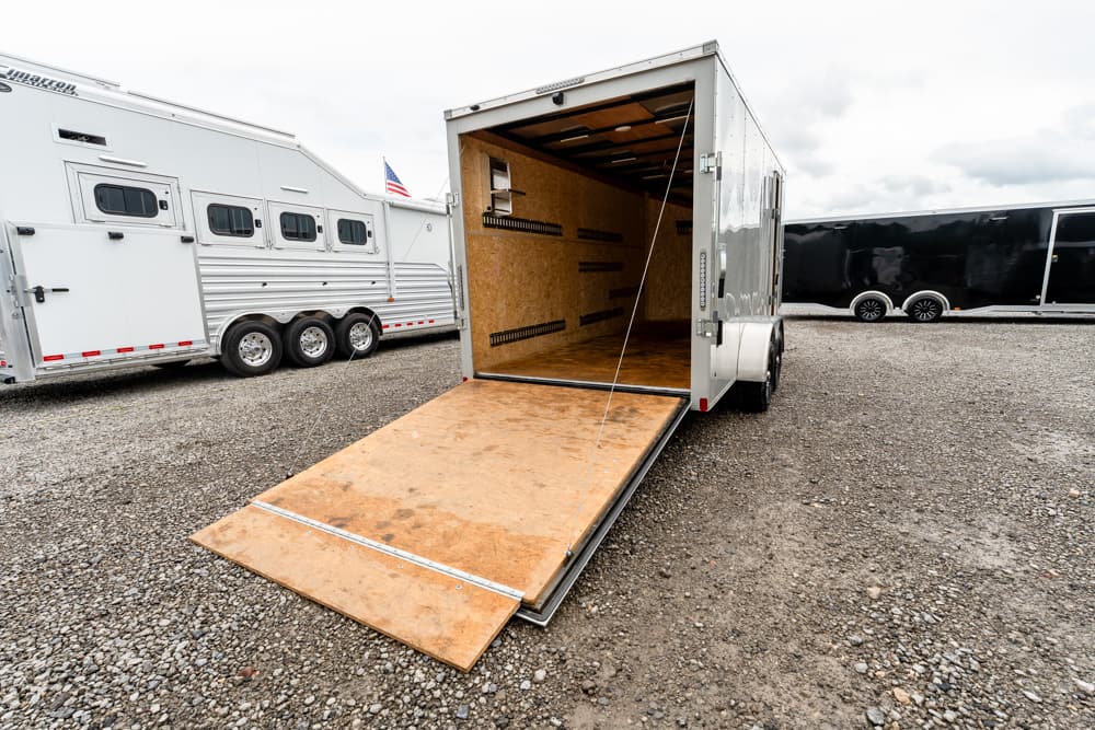 Open white cargo trailer with its wooden ramp lowered to the gravel, showing a plywood interior and tie-down rails; other trailers line the lot nearby.