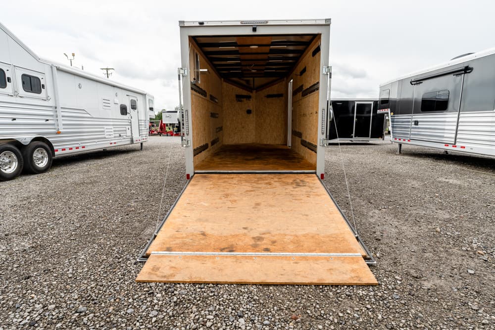 Open empty cargo trailer with a wooden ramp lowered for loading, parked on a gravel lot with other trailers nearby behind.
