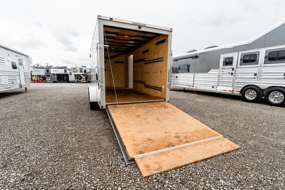Open cargo trailer with ramp down, empty interior, parked on a gravel lot among other show trailers.