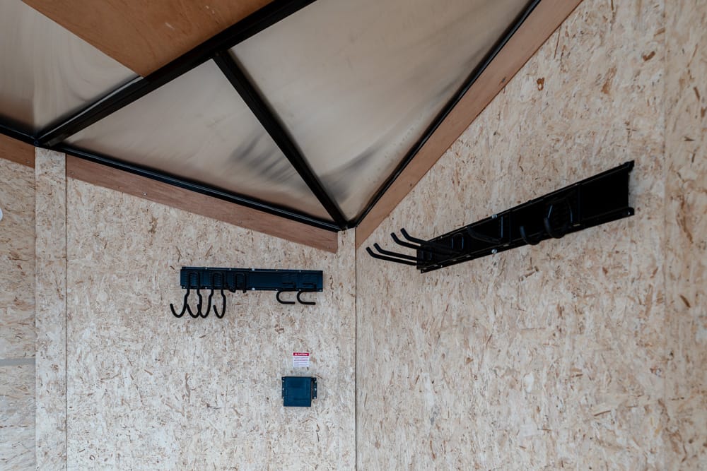 Indoor corner of a wooden shed with black metal coat hooks on plywood walls and a translucent sloped roof panel a overhead.