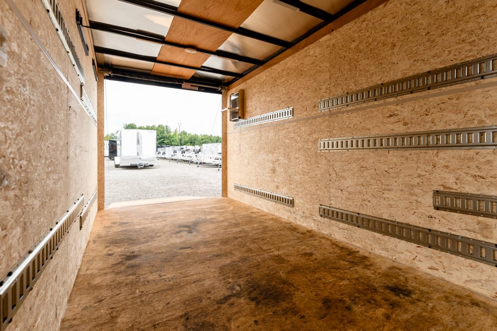 Interior of an empty cargo trailer with wooden walls and metal track rails, looking toward the open yard outside.