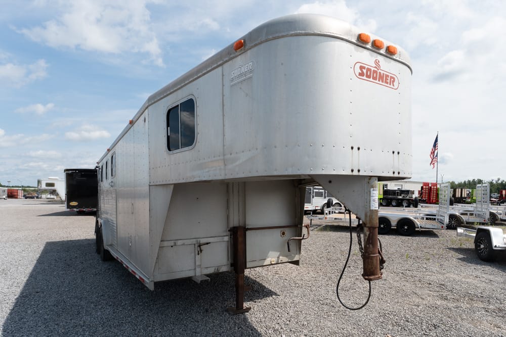 White gooseneck horse trailer parked on a gravel lot, showing a rusted hitch and small window on a sunny day with other trailers nearby.