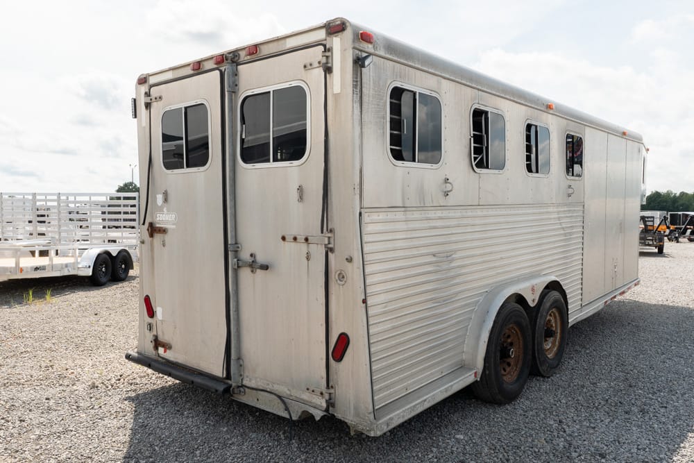 Silver livestock trailer on a gravel lot, showing rear double doors and small windows along the side.