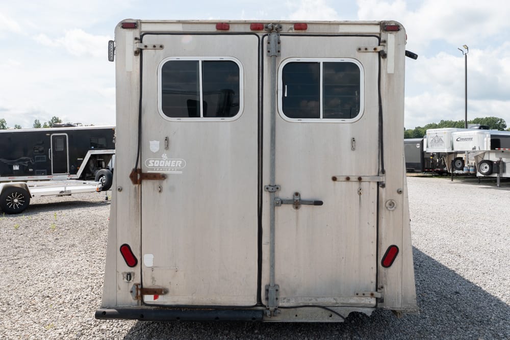 Rear view of a weathered two-door horse trailer with small windows and metal latches in a gravel trailer yard.