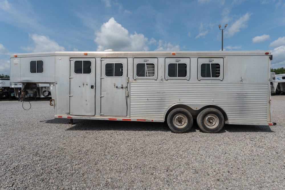 Silver aluminum horse trailer with multiple windows parked on a gravel lot, showing dual axles and rear and side doors under a blue sky.