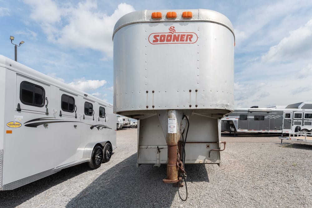 Front view of a large silver Sooner gooseneck trailer in a gravel lot, with white horse trailer/RVs on either side and a blue sky above.