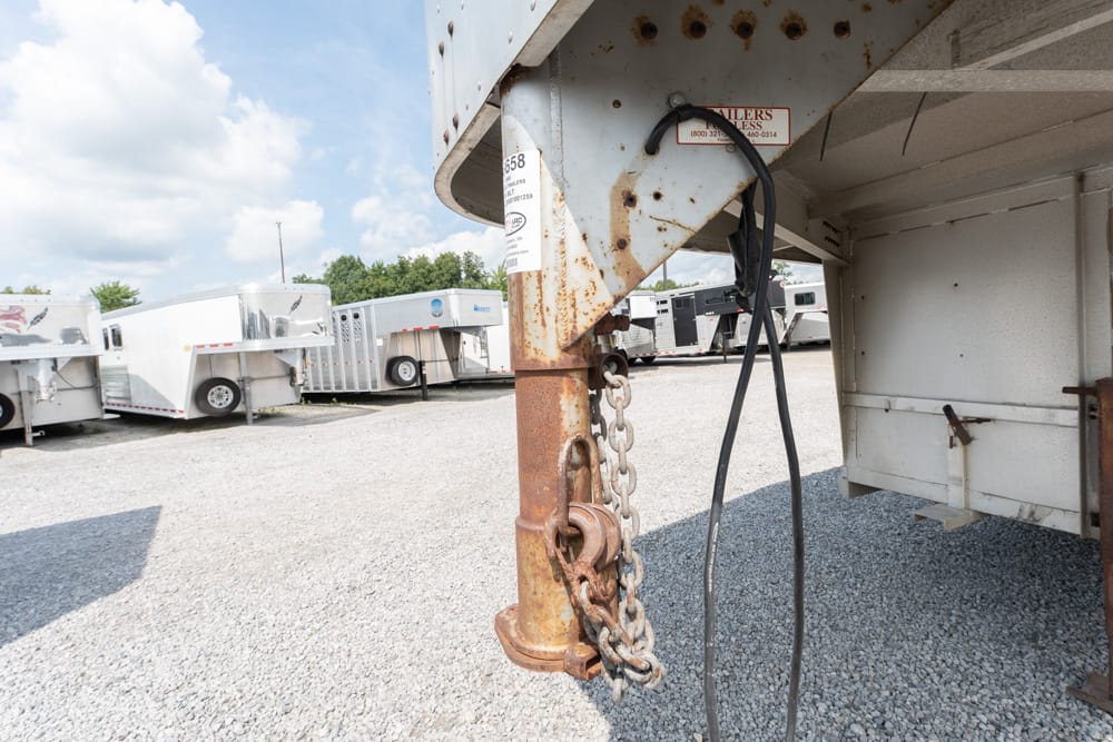 Close-up of a rusty trailer hitch and safety chains under a parked semi-trailer in a gravel lot.