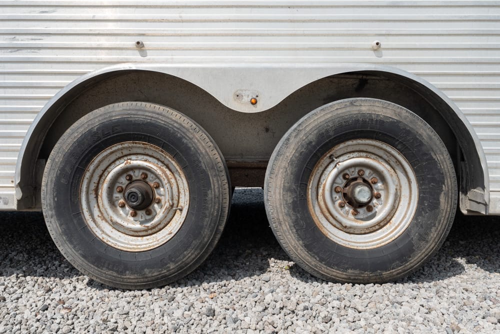 Two rusted trailer wheels side by side on a gravel surface beneath a metal trailer body with a small gap between them.
