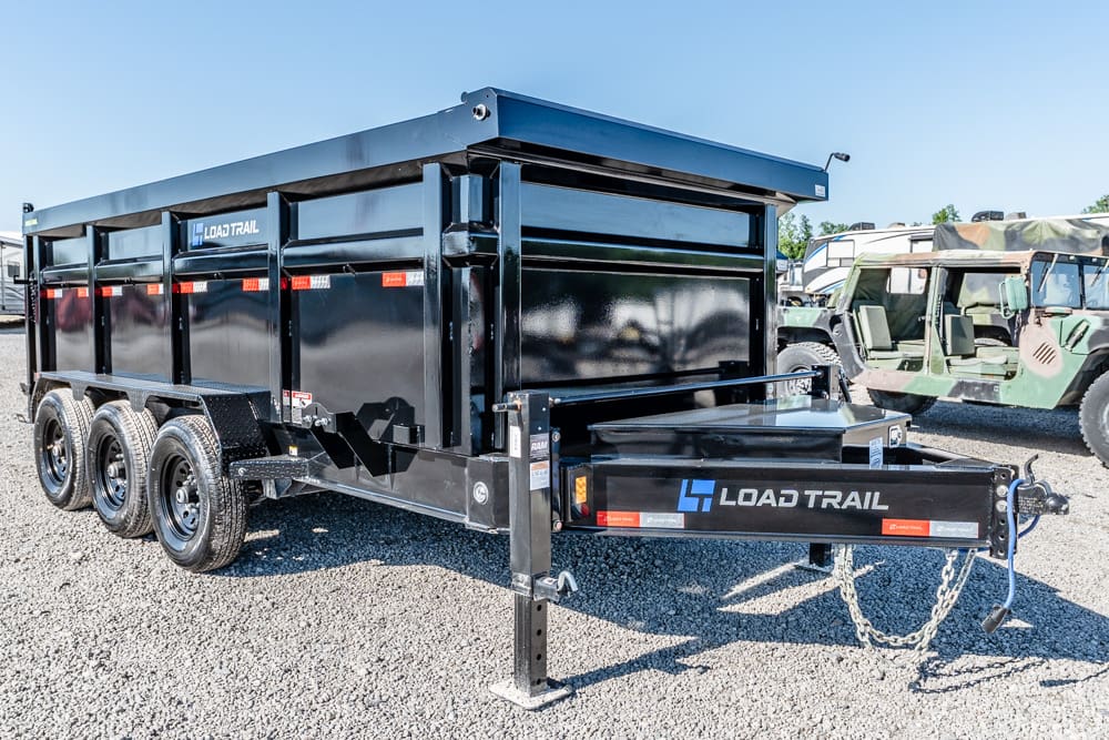 Black Load Trail dump trailer with three axles parked on gravel, hitch at front, in a lot with other vehicles nearby