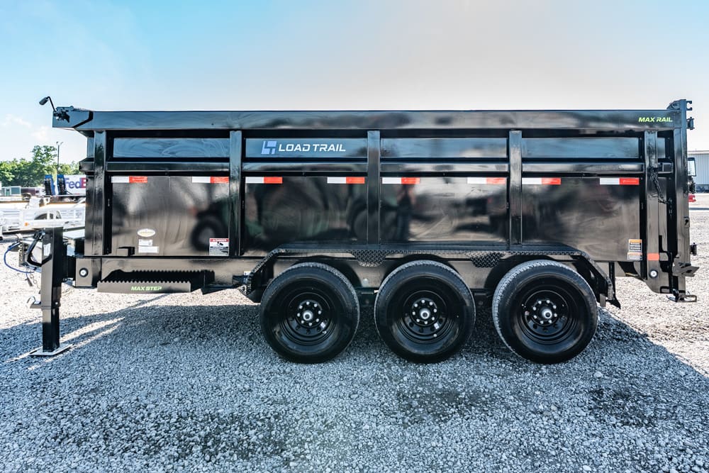 Black Load Trail dump trailer with three axles parked on a gravel lot, side view showing branding and reflective strips