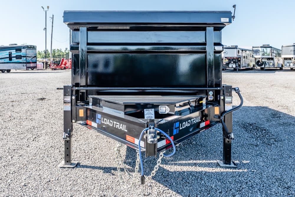 Front view of a black Load Trail dump trailer parked on gravel, showing the hitch, safety chains, and RVs in the background.