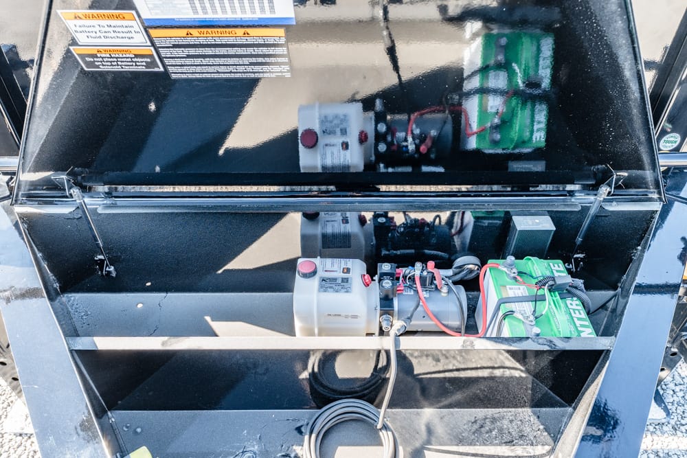 Open compartment of a portable generator showing battery, wiring, and a white reservoir with a red start button, inside a black metal housing.