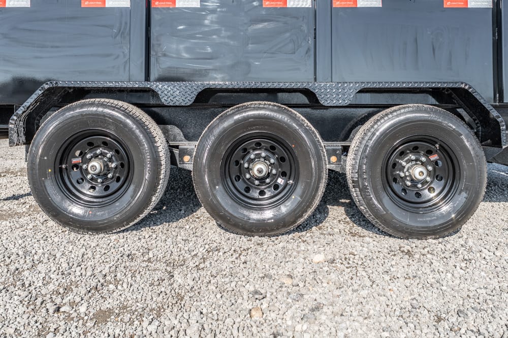 Triple-axle trailer wheel set with black rims on a gravel surface under a dark cargo trailer edge.