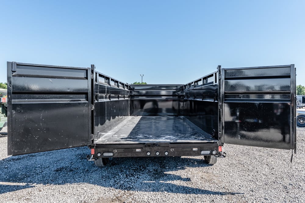 Back view of an empty black dump trailer with wide open rear doors on a gravel lot
