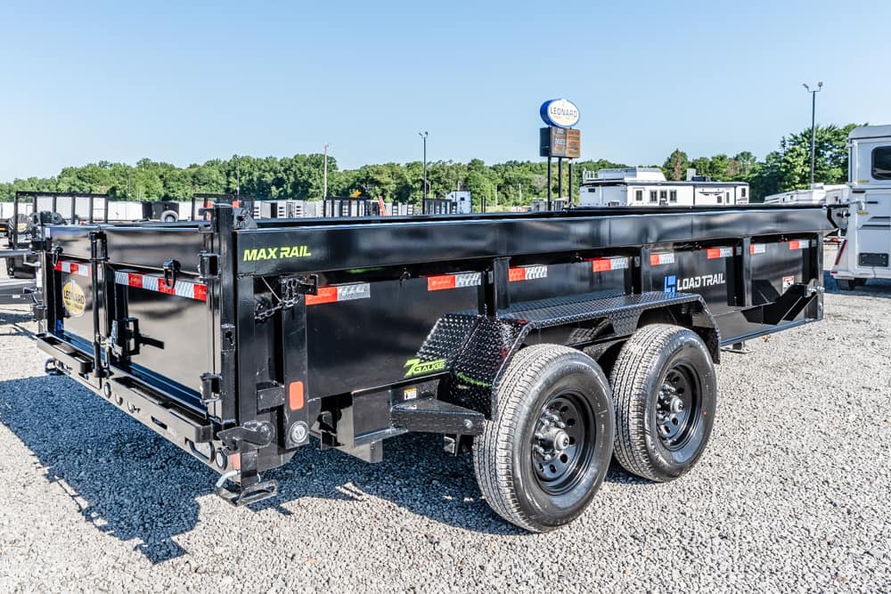 Black dual-axle dump trailer labeled MAX RAIL and LOAD TRAIL, parked on a gravel lot in a trailer yard.