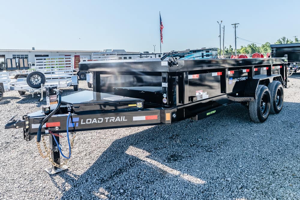 Black Load Trail dump trailer with dual axles and a raised bed on a gravel lot at a dealership. Tail section shows hitch, chains, and safety chains with roadside scene in background.