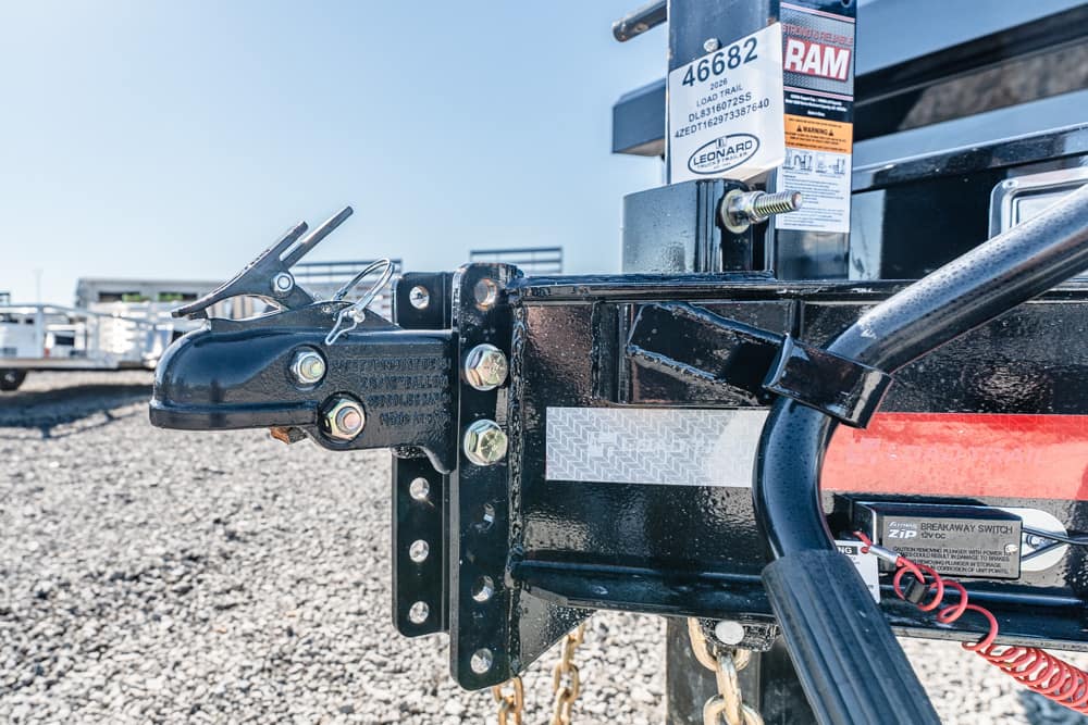 Close-up of a heavy-duty trailer hitch on a black trailer, showing bolts, safety chains, and labeling in a gravel lot