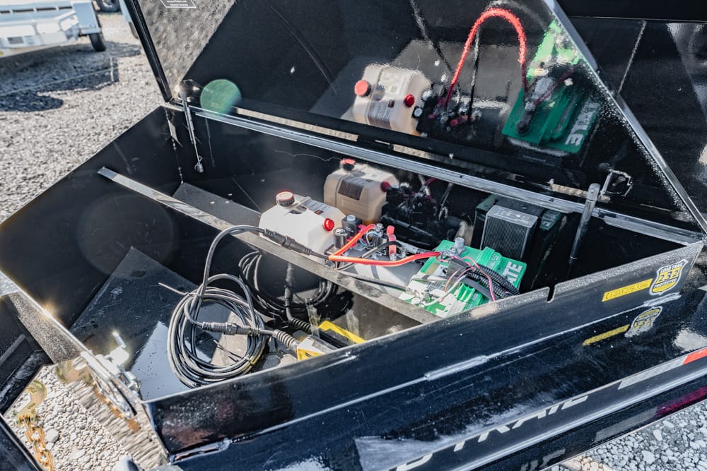 Open equipment box with batteries, wiring, and control boards in a black trailer box on a gravel surface